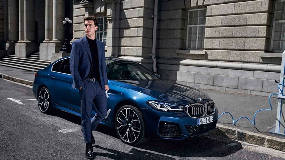 Mann in Anzug neben blauem BMW 5er Limousine vor einem Gebäude auf der Straße.