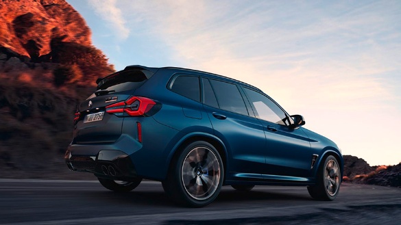 Blauer BMW X3 fährt auf Straße, im Hintergrund Berge bei Sonnenuntergang.