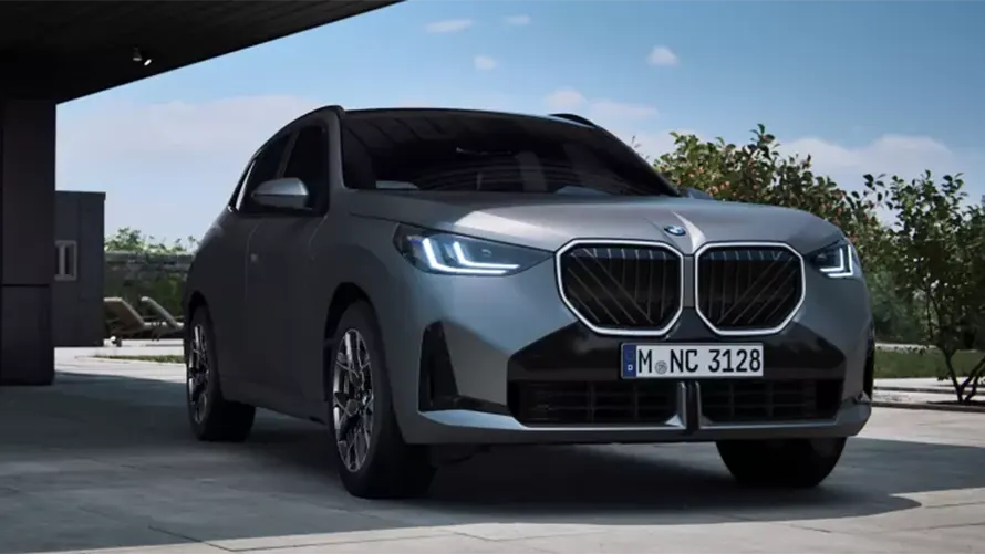 Silberner BMW SUV unter einem Vordach, Bäume im Hintergrund, sonniges Wetter.