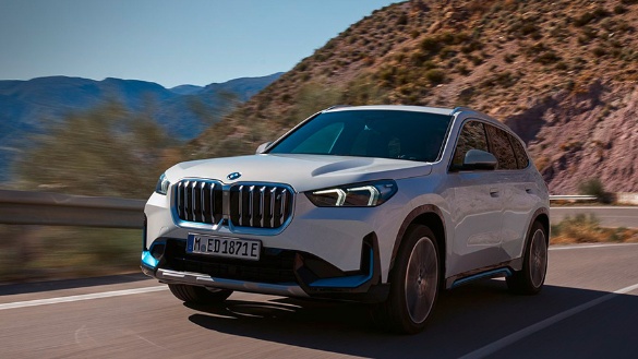 Weißer BMW iX1 fährt auf einer Straße in bergiger Landschaft bei Sonnenschein.