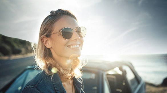 Frau mit Sonnenbrille lächelt vor einem Auto in sonniger Landschaft.