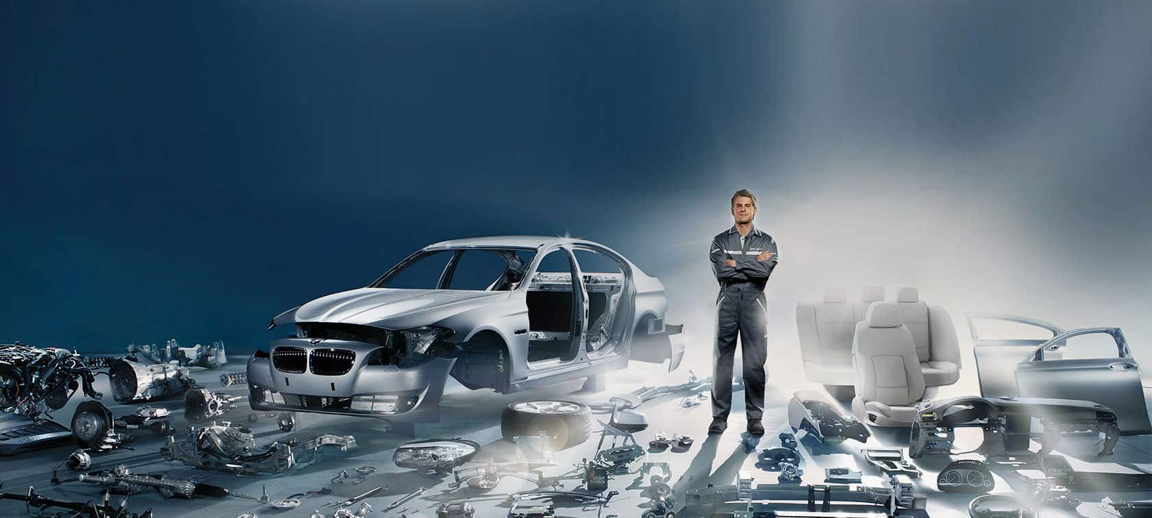 Ein Mechaniker steht vor einem teilweise zerlegten Auto mit Autoteilen um ihn herum.