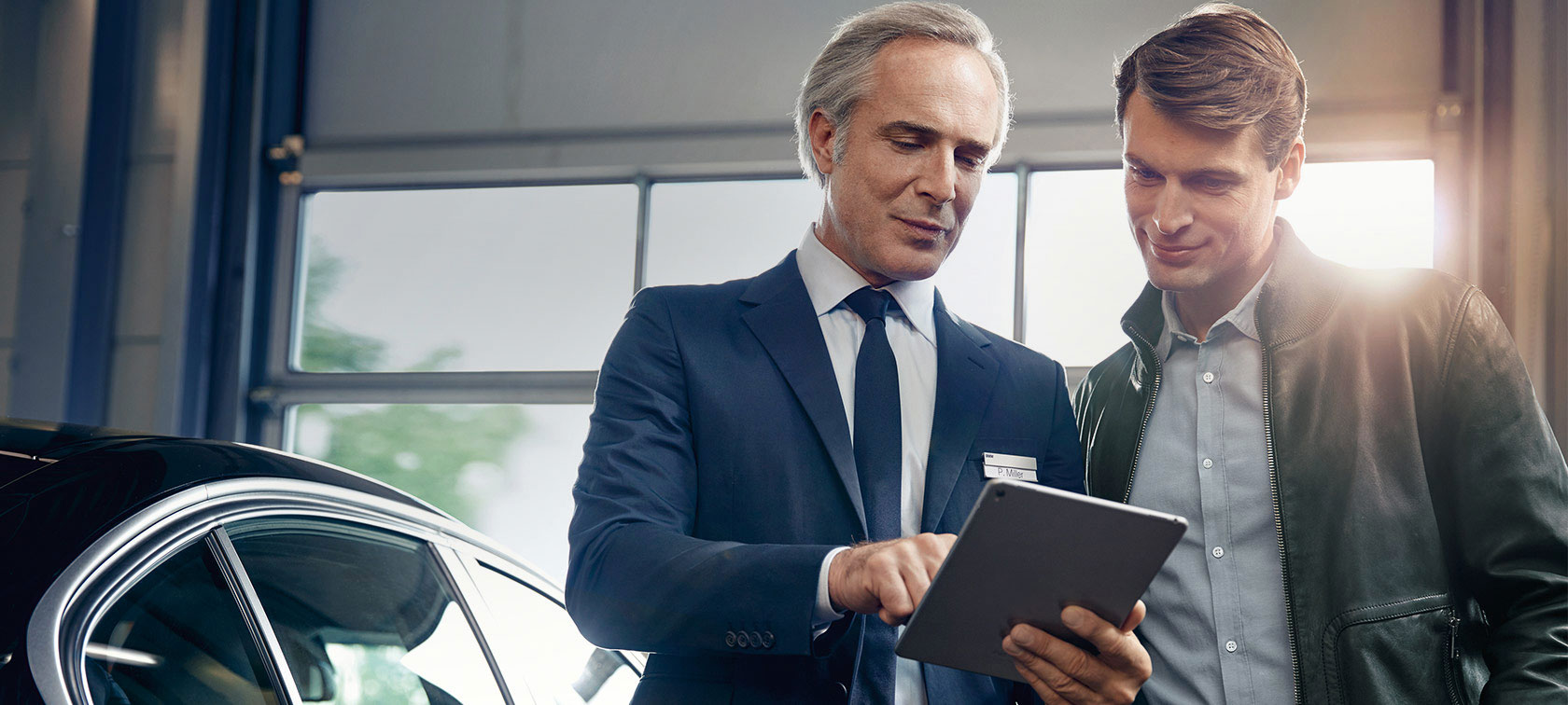 Zwei Männer in einem Autohaus, einer zeigt auf ein Tablet, ein Auto ist teilweise sichtbar.