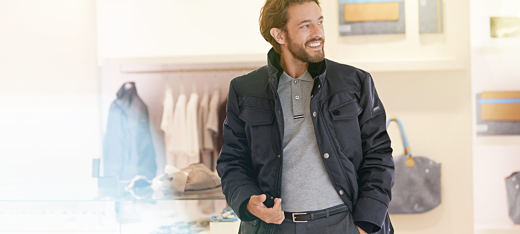 Mann in schwarzer Jacke und grauem Hemd lächelt, im Hintergrund helle Kleidung und Taschen.