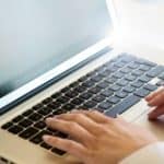 Hände tippen auf einer Laptop-Tastatur. Bildschirm ist teilweise sichtbar.