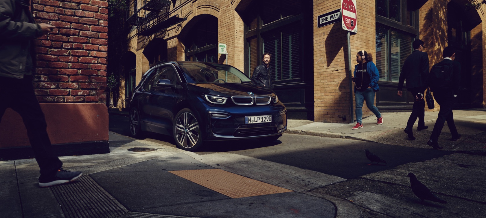 Ein schwarzer BMW i3 parkt in einer urbanen Straße mit Passanten und Ziegelgebäuden.