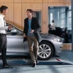 Zwei Männer stehen lächelnd vor einem silbernen Auto in einem Showroom.