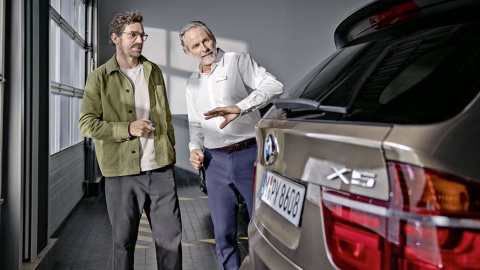 Zwei Männer stehen neben einem braunen BMW X5 in einem hellen Raum, einer erklärt etwas.
