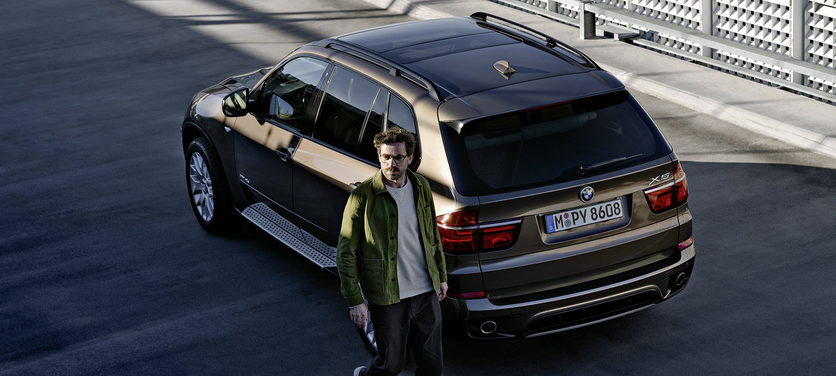 Ein Mann in grüner Jacke steht neben einem braunen BMW X5 auf einem Parkplatz.