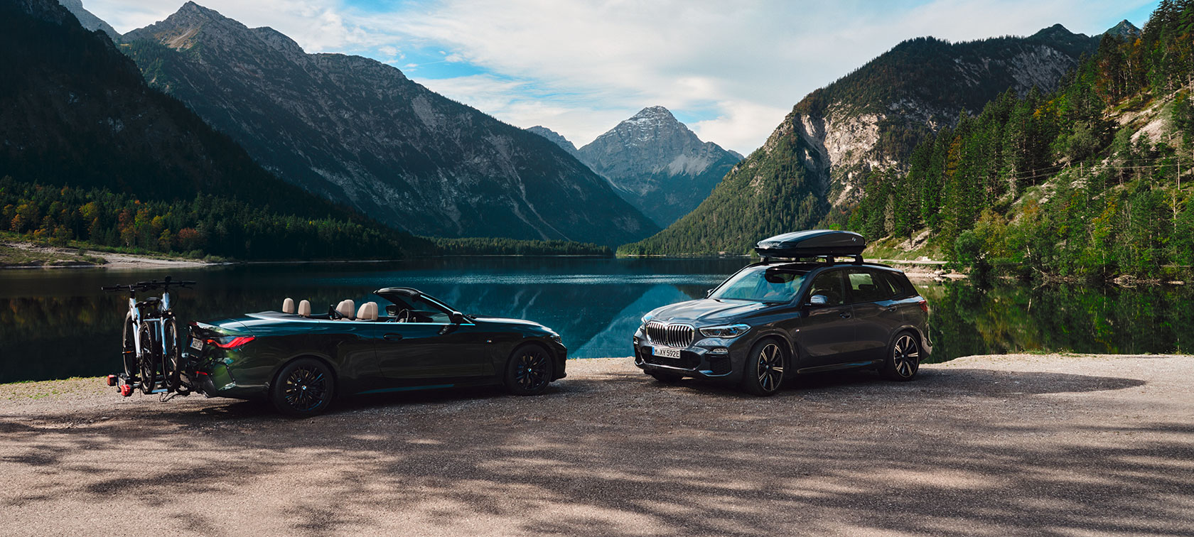 Zwei BMW-Fahrzeuge am Seeufer, umgeben von Bergen und Wald, bei klarem Himmel.