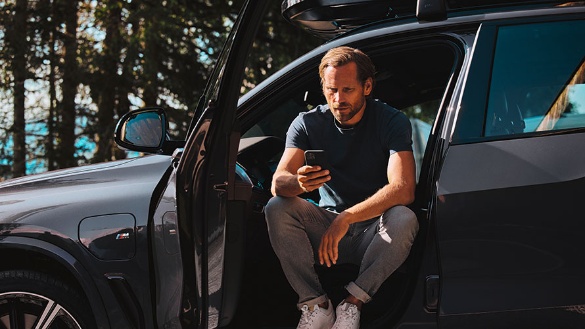 Mann sitzt in geöffneter Autotür eines grauen BMW und schaut auf sein Smartphone.