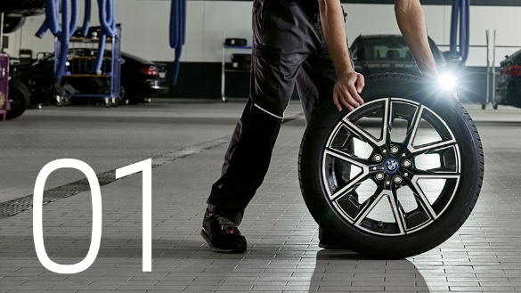 Person in Werkstatt zieht BMW-Reifen mit Felge, Zahl 01 links.