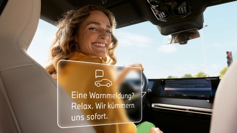 Frau im Auto, lächelt zur Seite, Display mit BMW-Warnmeldung, sonniger Himmel im Hintergrund.