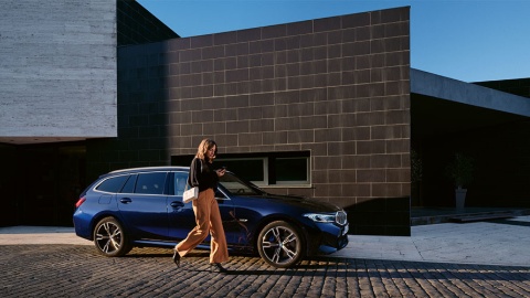 Frau geht an einem blauen BMW vorbei, modernes Gebäude im Hintergrund, bei Sonnenuntergang.