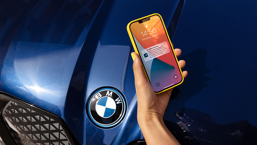 Hand hält gelbes Smartphone mit Benachrichtigung über BMW-Logo auf blauem Autolack.