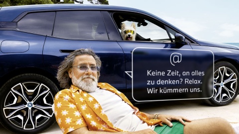 Älterer Mann in Hawaiihemd vor blauem BMW, Hund im Auto, Texttafel mit Entspannungsbotschaft.