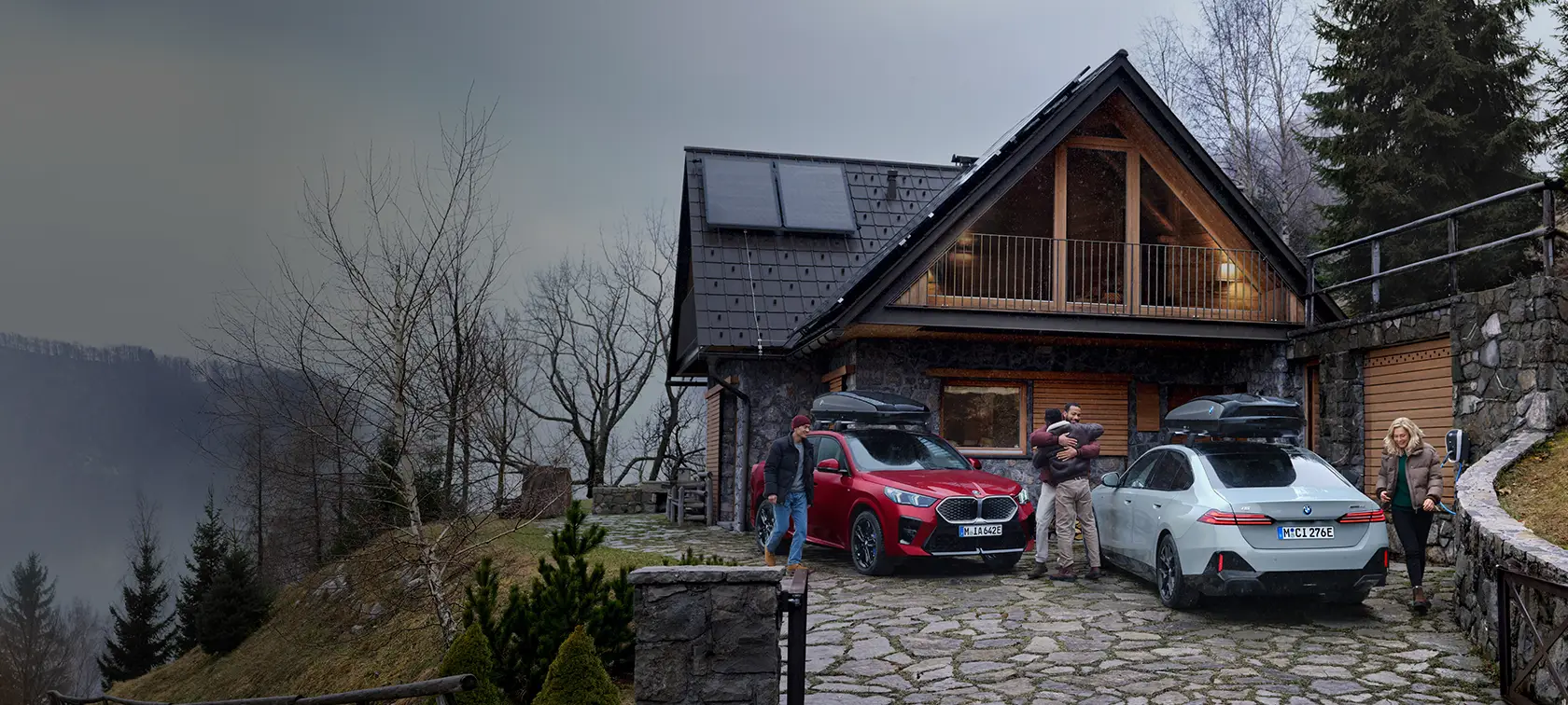 Zwei geparkte BMWs vor einem Haus im Wald, Personen umarmen sich und laden ein Auto auf.