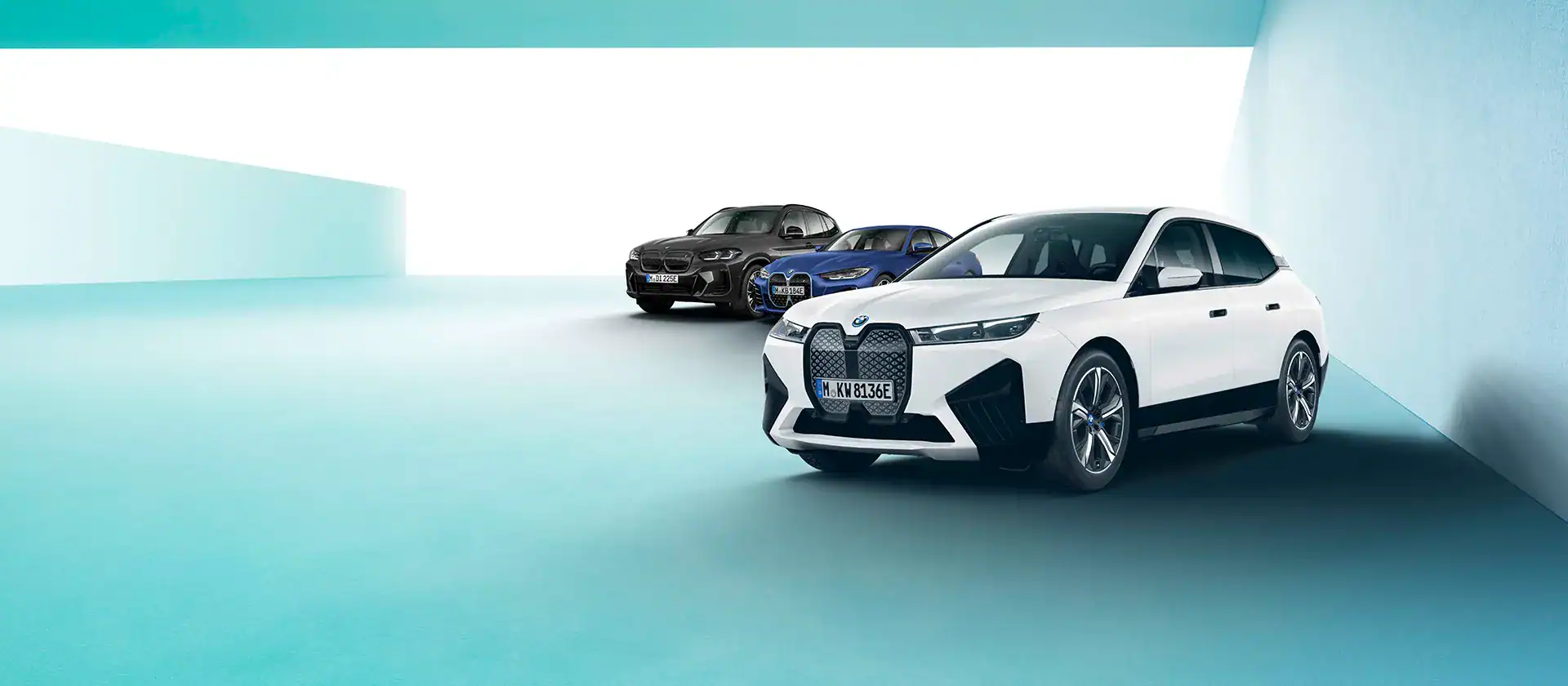 Drei BMW-Fahrzeuge in hellem Raum, vorne weißer SUV, dahinter schwarze und blaue Limousinen.