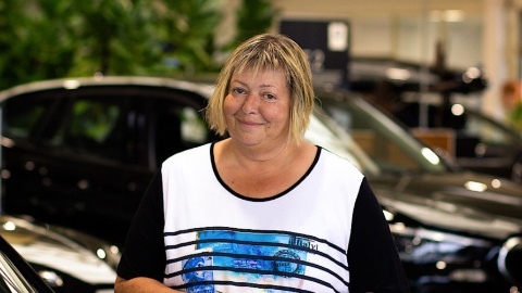 Person in einem Autohaus, lächelnd, mit Autos im Hintergrund.