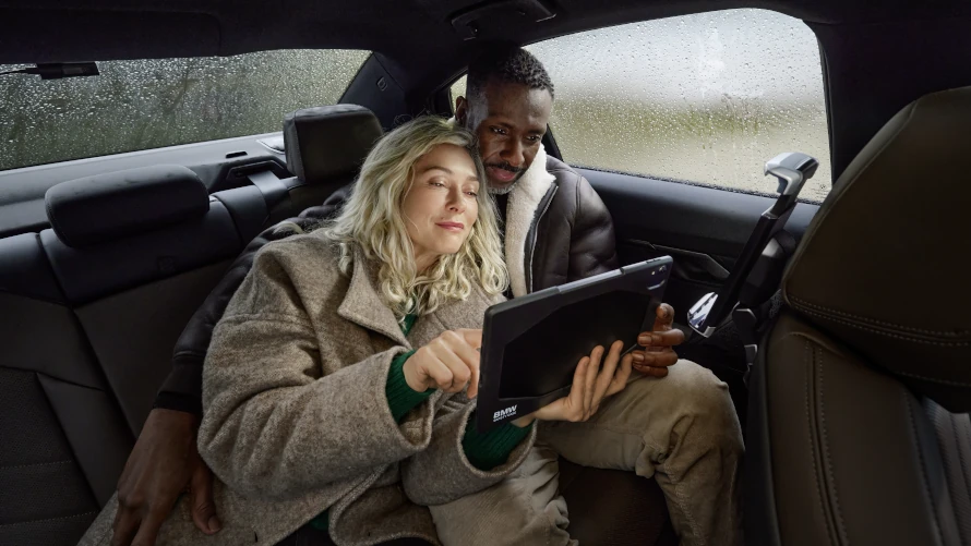 Paar sitzt auf Rücksitz eines Autos und hält ein Tablet mit BMW-Logo, draußen regnet es.