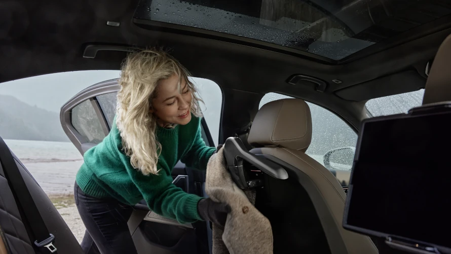 Frau in grünem Pullover hängt Mantel an Kleiderhalter im Auto auf, See im Hintergrund.