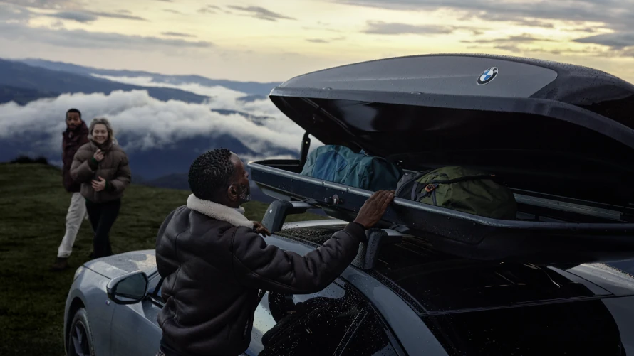 Person öffnet BMW-Dachbox am Auto in Berglandschaft; zwei Menschen im Hintergrund.