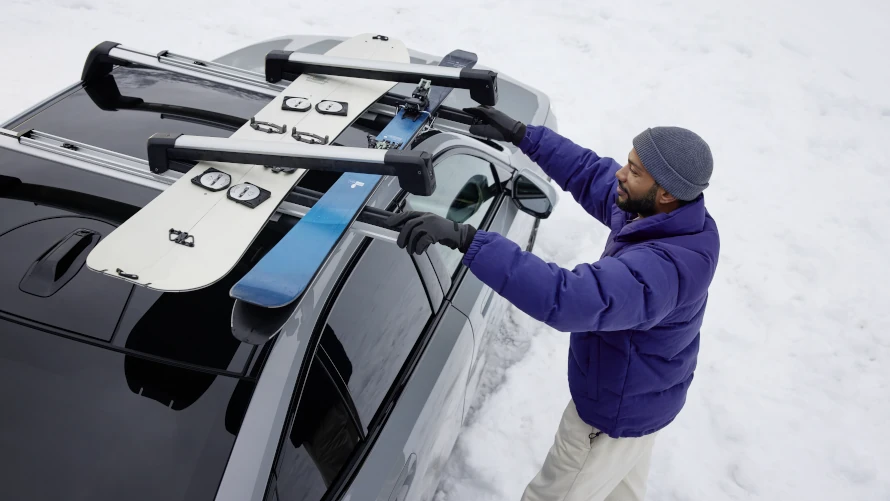 Person befestigt Ski auf Dachträger eines schwarzen Autos im Schnee.