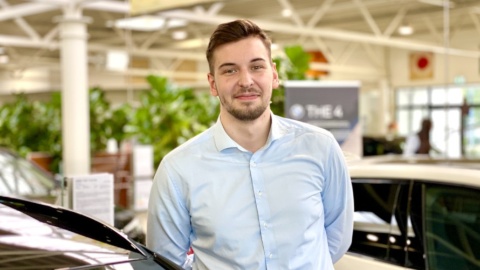 Mann in hellblauem Hemd in einem Autohaus, umgeben von Pflanzen und Fahrzeugen.