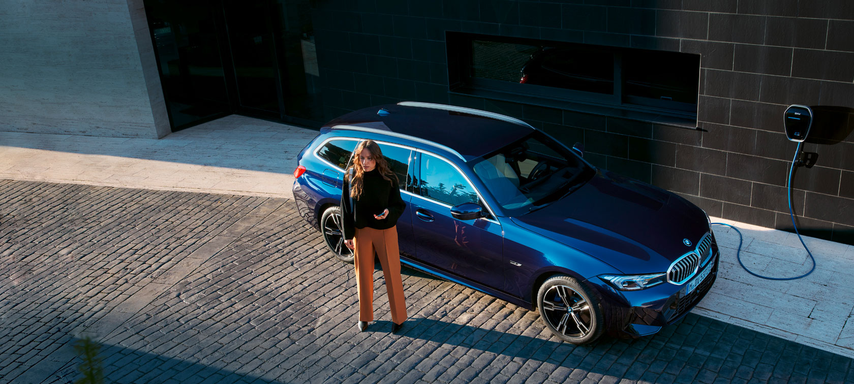 Frau steht neben einem blauen BMW, der an einer Ladestation angeschlossen ist, auf gepflastertem Boden.