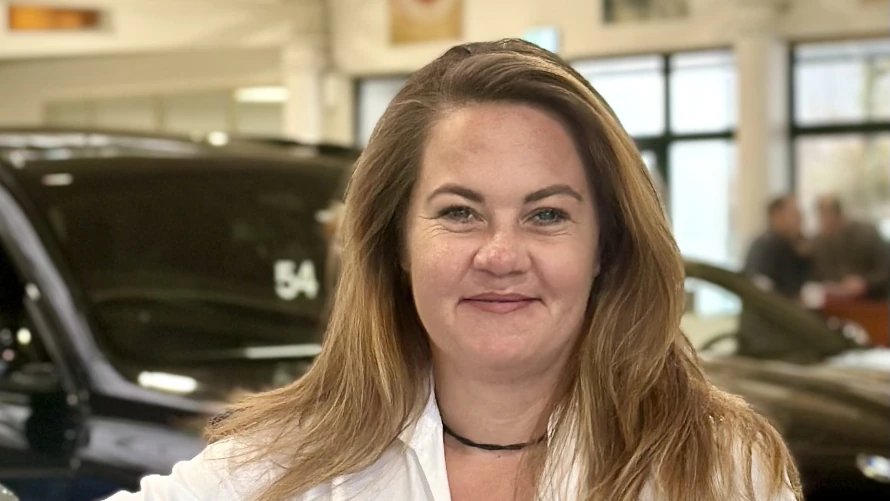 Eine lächelnde Frau in einem Autohaus mit Autos im Hintergrund.