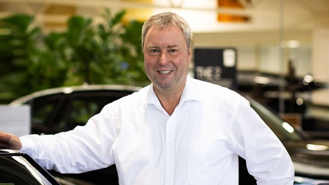 Ein Mann in weißem Hemd steht lächelnd in einem Autohaus neben einem schwarzen Auto.