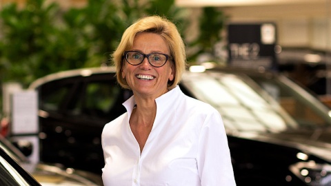 Frau mit Brille und weißem Hemd lächelt in einem Autohaus, im Hintergrund schwarze Autos.