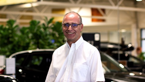 Ein lächelnder Mann mit Brille in einem Autohaus, umgeben von Autos und Pflanzen.