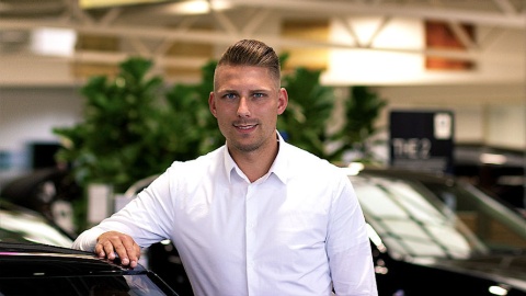 Mann im weißen Hemd lehnt an einem Auto in einem Autohaus mit unscharfem Hintergrund.