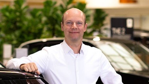 Ein Mann in weißem Hemd steht lächelnd neben einem Auto in einem Ausstellungsraum.