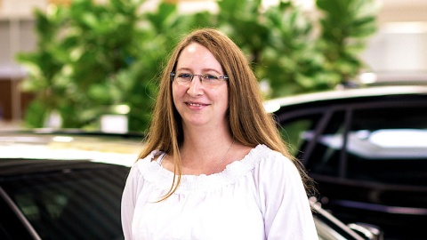 Eine lächelnde Frau mit Brille steht vor einem Auto in einem Ausstellungsraum.