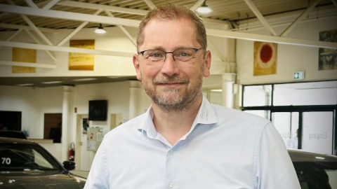 Ein Mann mit Brille steht lächelnd in einem Autohaus neben einem schwarzen Auto.