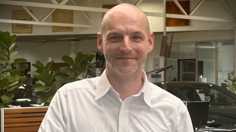 Ein Mann in weißem Hemd lächelt in einem Autohaus mit Pflanzen im Hintergrund.