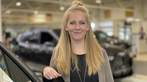 Eine Person mit langen, blonden Haaren steht in einem Autohaus, lächelt in die Kamera.