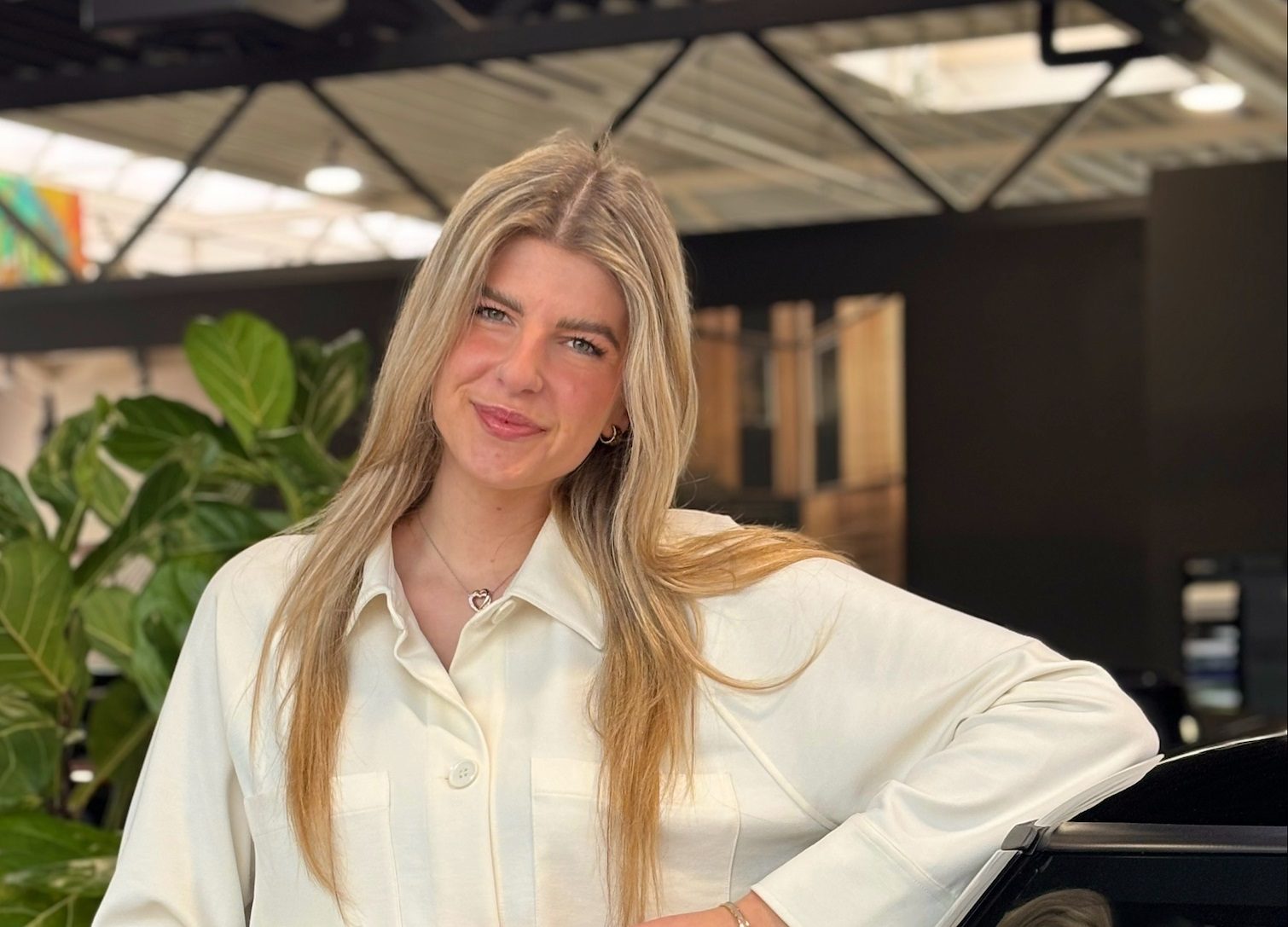 Eine Frau in weißem Hemd lächelt, lehnt an einem Auto, Pflanzen im Hintergrund.