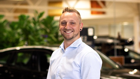 Eine lächelnde Person in einem Autohaus mit Pflanzen und Fahrzeugen im Hintergrund.