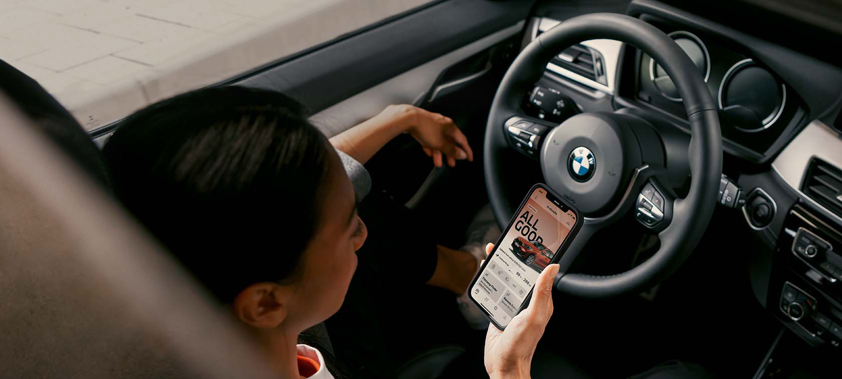Person im Auto mit Smartphone, BMW-Logo auf Lenkrad sichtbar.