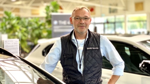 Ein Mann mit Brille und BMW-Weste steht in einem Autohaus neben einem weißen Auto.
