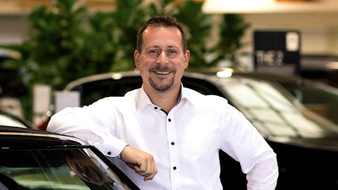 Mann in weißem Hemd lehnt an einem Auto, im Hintergrund Pflanzen und Autos in einem Showroom.