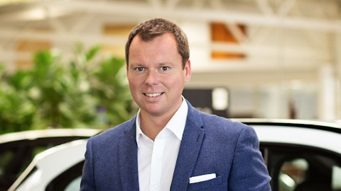 Mann in blauem Anzug vor einem weißen Auto in einem Autohaus.