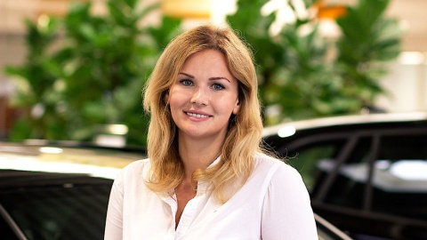 Eine lächelnde Frau in weißer Bluse steht vor unscharfem Hintergrund mit Pflanzen.