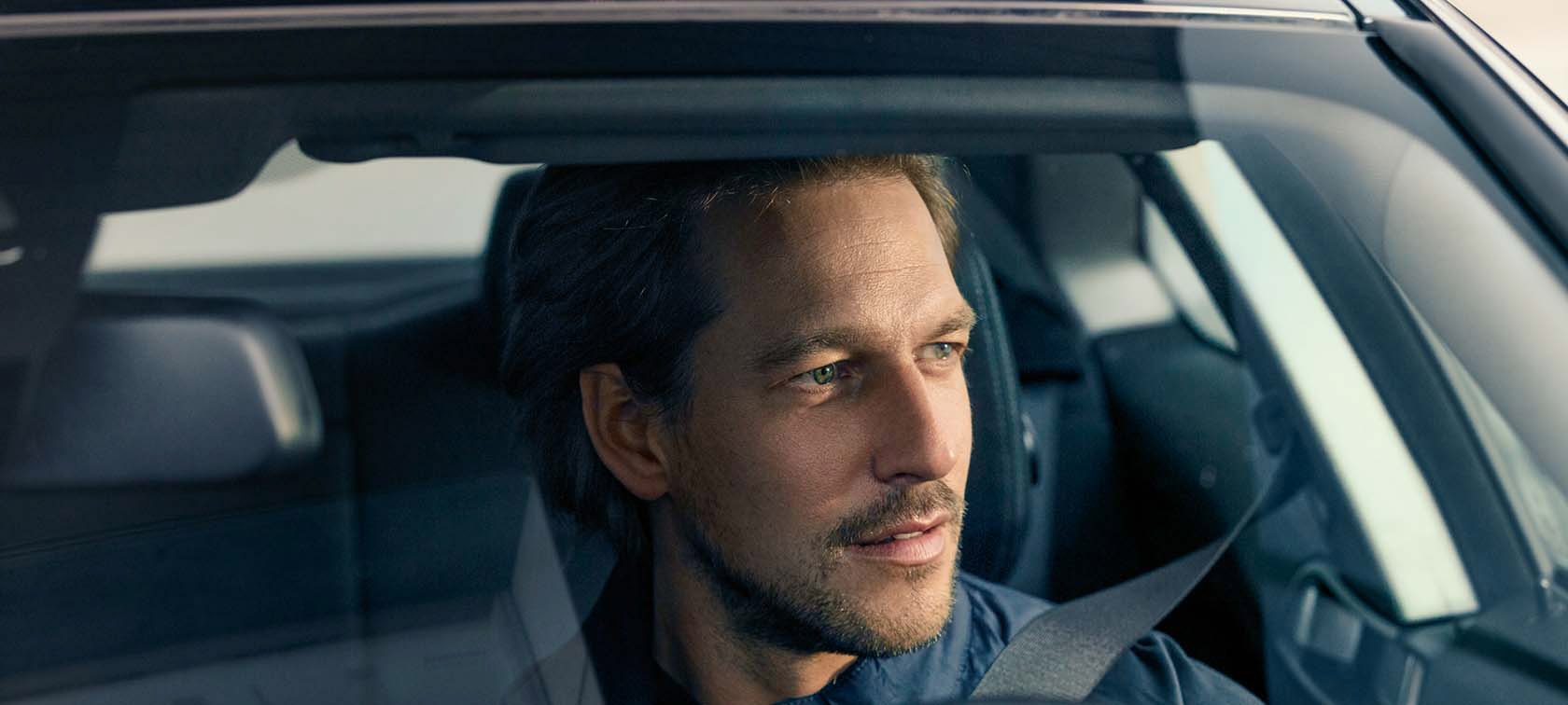 Mann sitzt in einem Auto, schaut aus dem Fenster, trägt Sicherheitsgurt, Innenraum ist sichtbar.