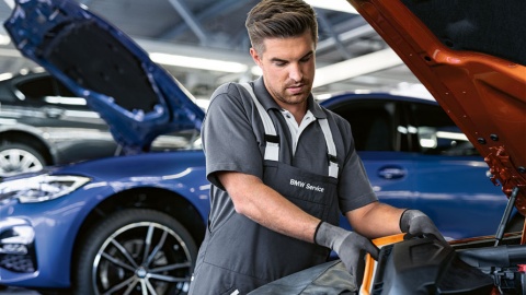 Mechaniker arbeitet am Motorraum eines orangefarbenen Autos, blauer BMW im Hintergrund.