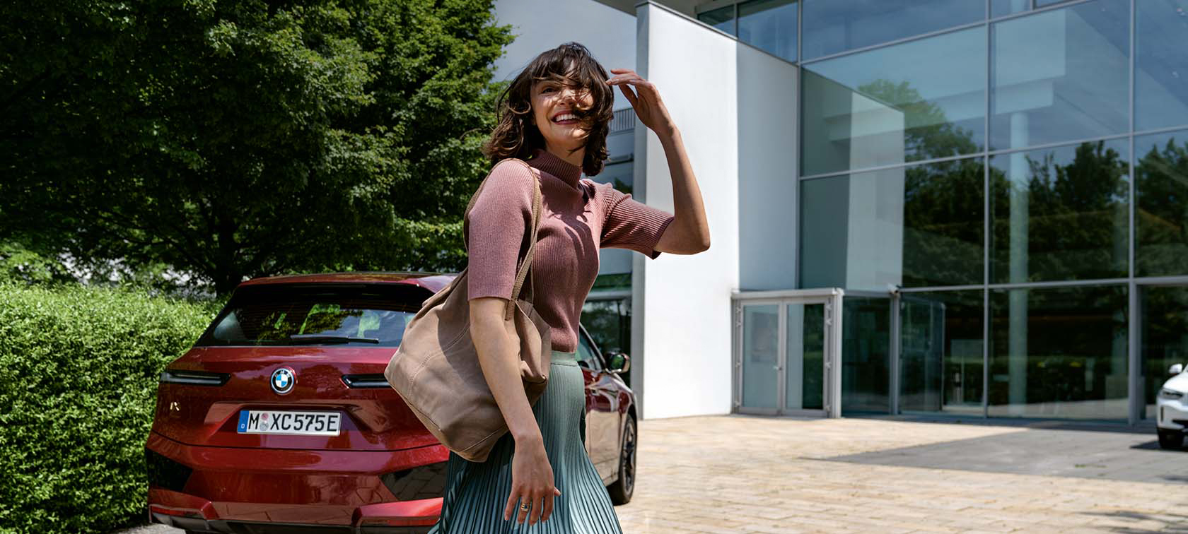 Frau vor rotem BMW iX, lächelnd, trägt rosa Oberteil, Tasche, steht vor einem modernen Gebäude.