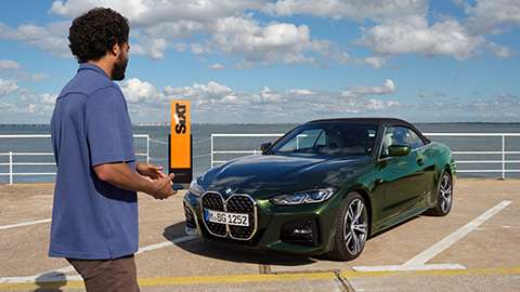 Ein Mann betrachtet ein grünes BMW Cabrio auf einem Parkplatz am Wasser bei Sonnenschein.
